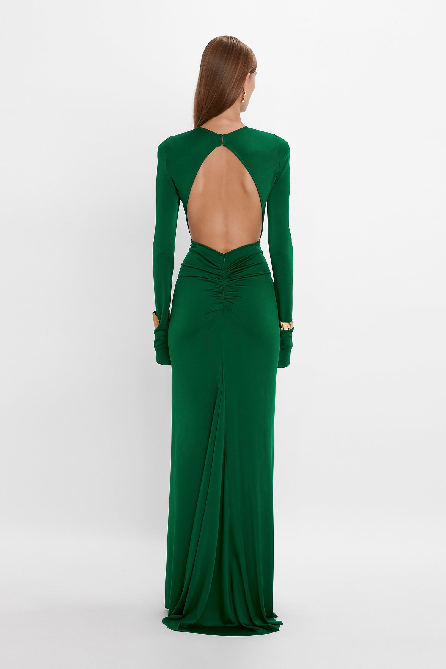 A woman stands facing away, wearing the Circle Detail Open Back Gown In Emerald by Victoria Beckham. Her hair is straight and falls down her back. The longer-length hem gathers at the lower back and extends to the floor, showcasing a body-sculpting jersey that enhances her silhouette effortlessly.