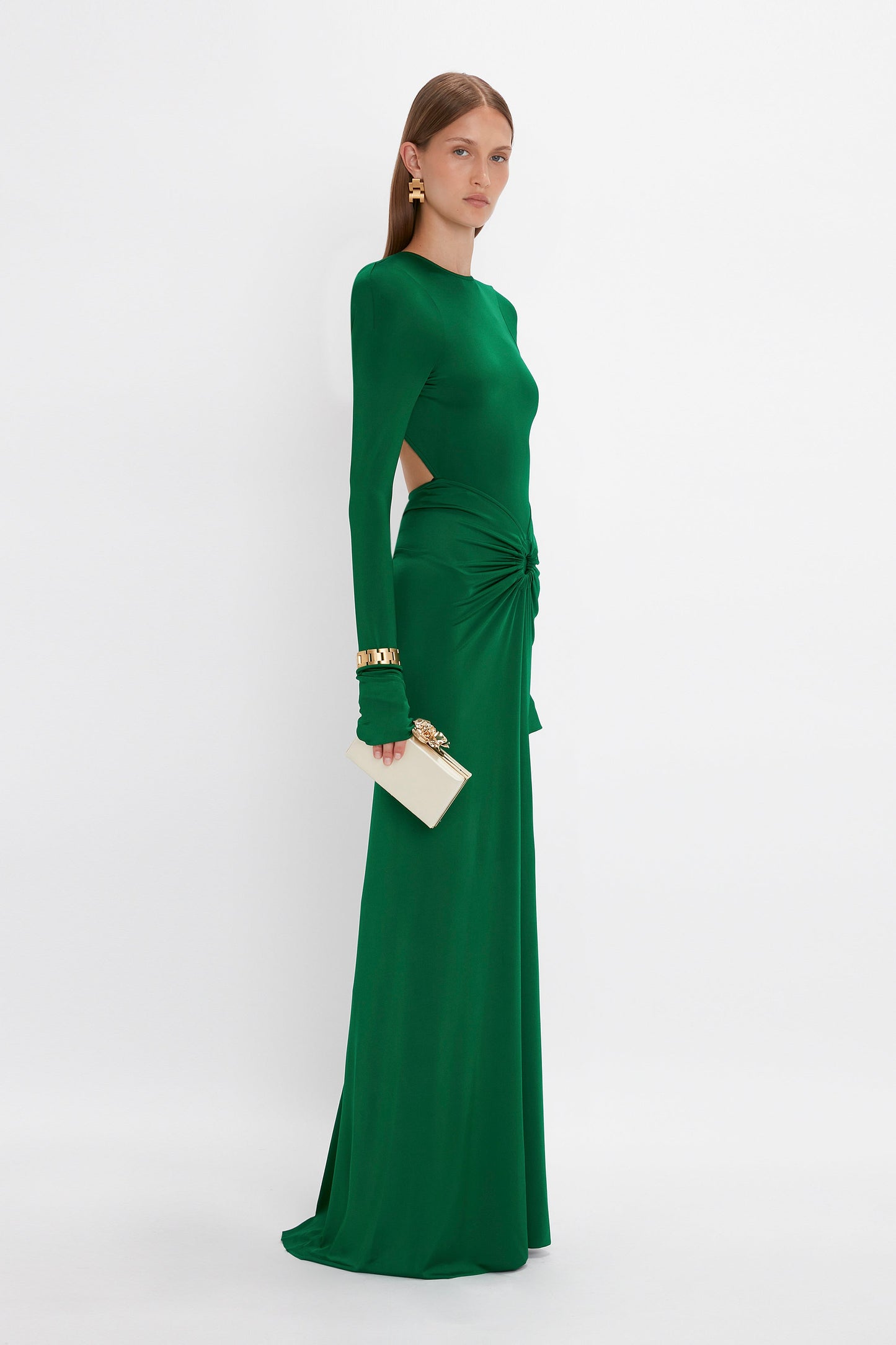 Woman in a **Victoria Beckham** **Circle Detail Open Back Gown In Emerald**, featuring side cut-outs and a knotted detail at the waist, with a longer-length hem. She holds a white clutch purse and accessorizes with gold earrings and a bracelet.