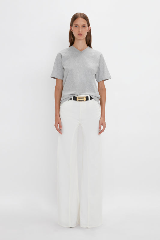 Person standing against a white background, wearing a Victoria Beckham Football T-Shirt In Grey Marl, white high-waisted wide-leg pants, and a black belt with a prominent gold buckle.