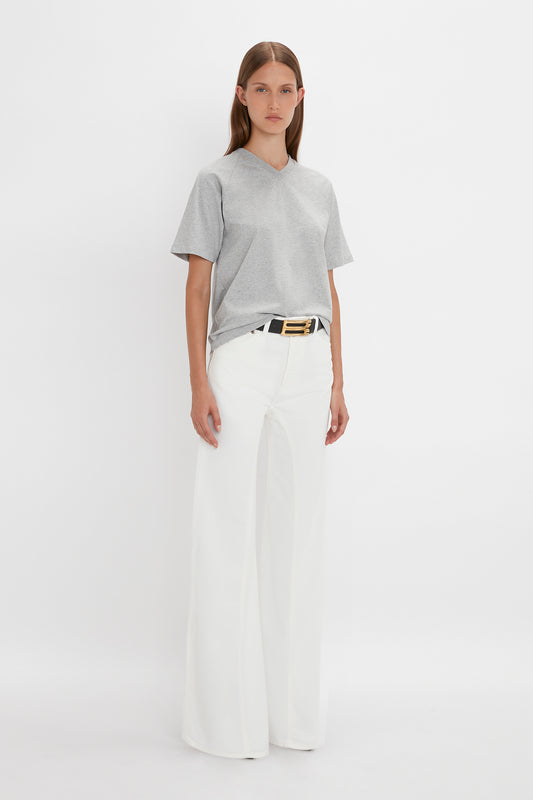 A person stands against a blank background wearing a Victoria Beckham Football T-Shirt in Grey Marl, white wide-leg pants, and a black belt with a gold buckle.
