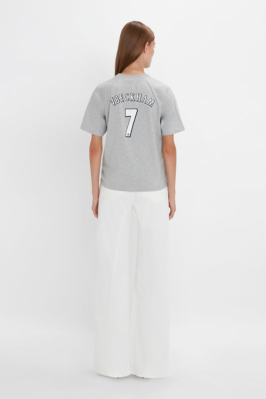 A person with long hair, standing and facing away, is wearing a Victoria Beckham Football T-Shirt In Grey Marl with the name "V. Beckman" and the number "7" on the back, paired with white pants. The background is plain white.