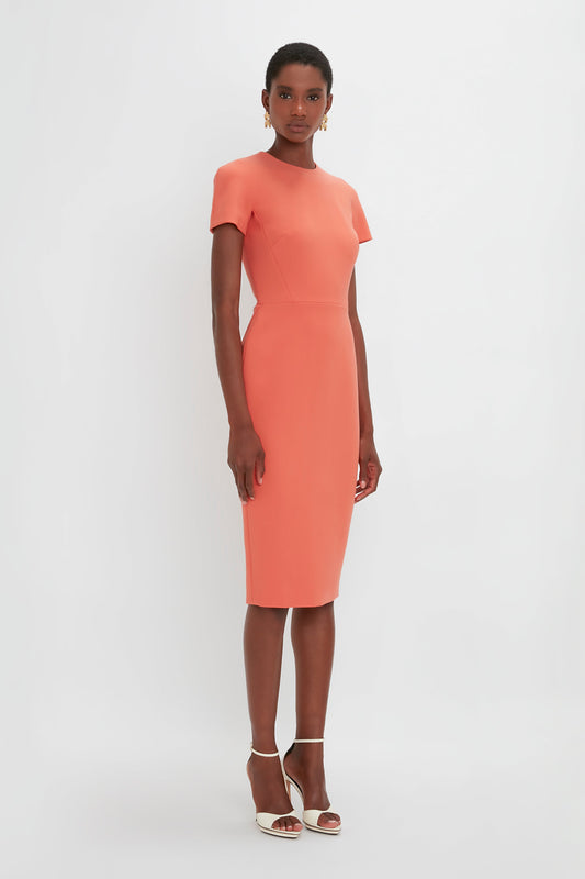 A woman standing straight in a studio, wearing an elegant coral midi-length dress with short sleeves and high heels by Victoria Beckham.