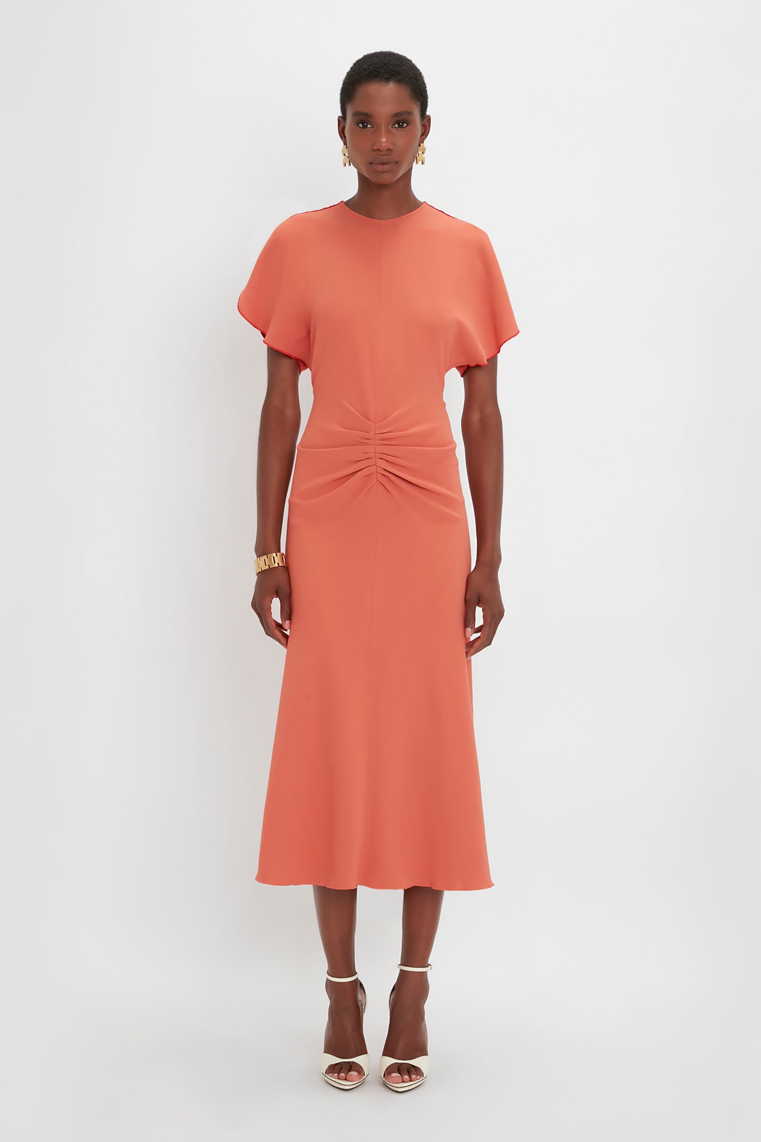 A woman standing against a white background, wearing a Victoria Beckham gathered waist midi dress in papaya with short sleeves and a twist detail at the waist. She has on pointy toe stiletto sandals and a watch.