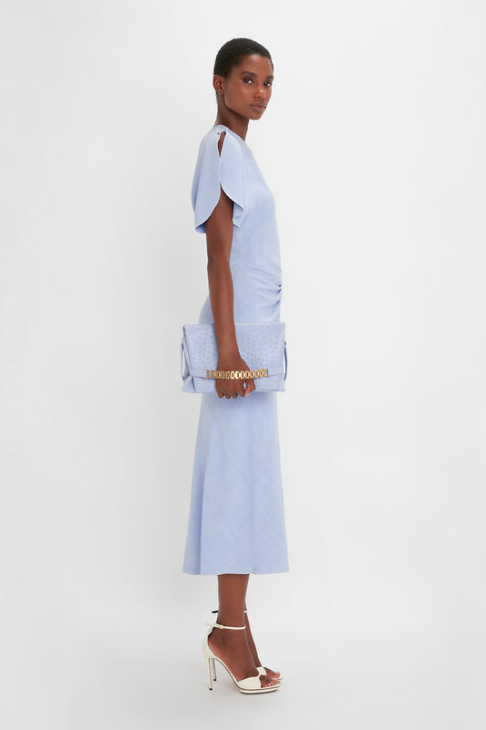 A person models a light blue, short-sleeve, knee-length dress paired with white high-heeled sandals. The individual carries a matching light blue clutch bag adorned with a subtle gold chain detail: the Chain Pouch With Strap In Frost Ostrich-Effect Leather by Victoria Beckham.