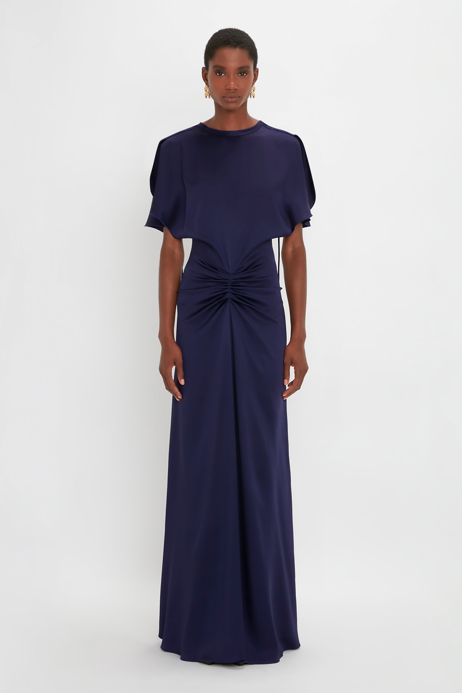 A person stands against a white background wearing a floor-length, navy blue dress with a knotted detail at the waist and Victoria Beckham's Exclusive Jumbo Chain Earrings in Brushed Gold.