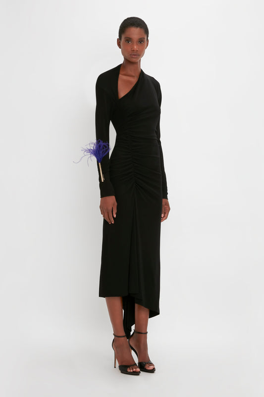 A woman models a Victoria Beckham black long-sleeve evening gown with a ruched waist and an asymmetric cut-out, paired with strappy heels against a white background.