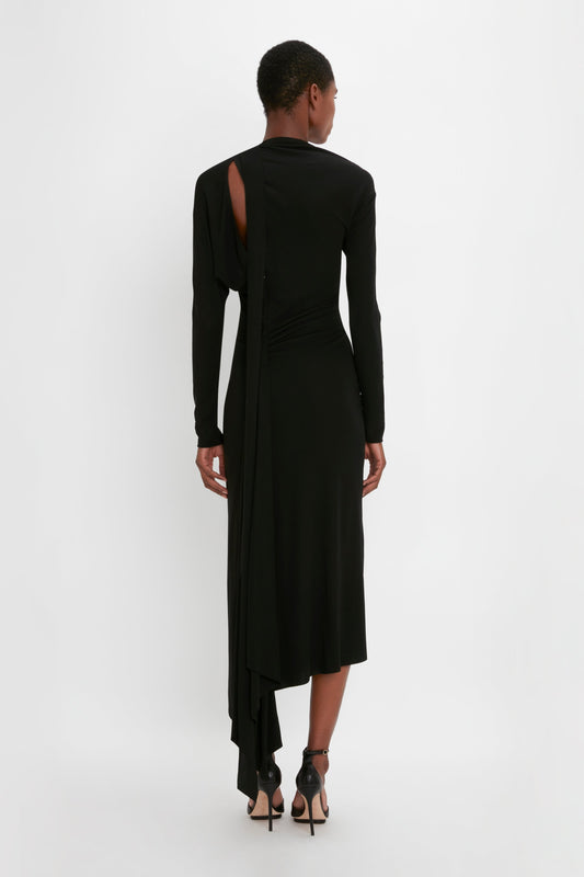 A woman in a Victoria Beckham body-sculpting black gown with an asymmetric cut-out and hem, viewed from behind, standing against a white background.