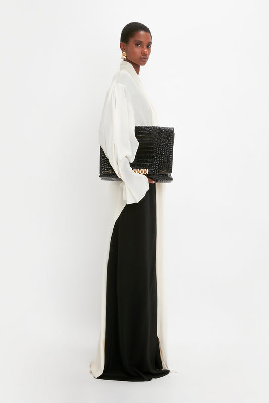 Person in a white blouse and black skirt holding a Victoria Beckham Jumbo Chain Pouch in Black Croc-Effect Leather, standing against a plain white background.