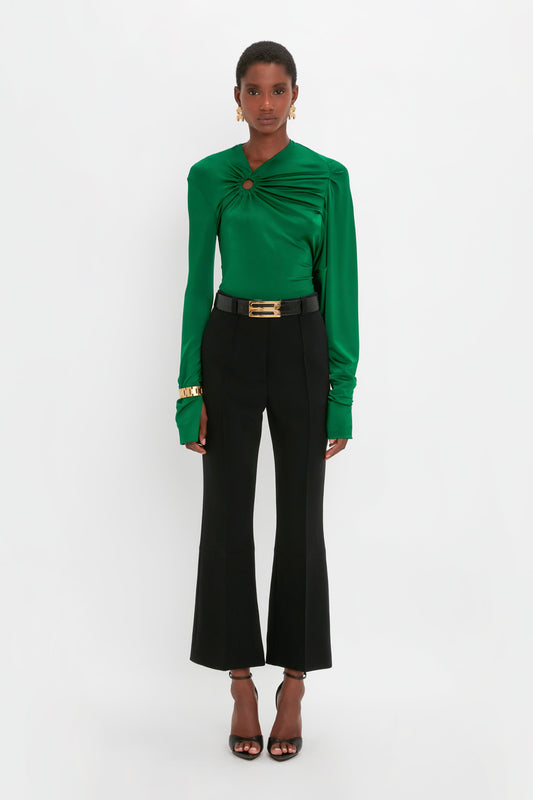 A person stands against a plain white background wearing a green, long-sleeve top paired with contemporary black Victoria Beckham Cropped Kick Trouser In Black and black heels.