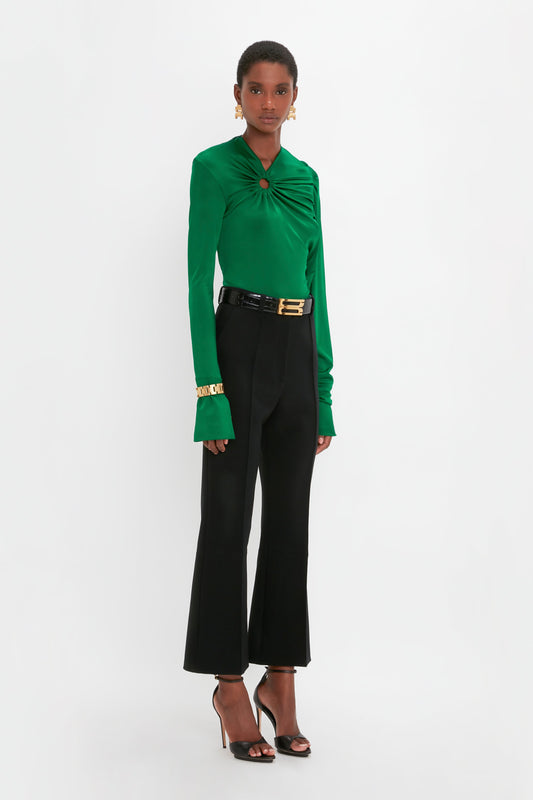 A woman stands against a plain background wearing a green long-sleeve top with a circular detail, Cropped Kick Trouser In Black by Victoria Beckham, black heeled sandals, a black belt with a gold buckle, and gold bracelets on her right wrist. This contemporary outfit perfectly blends elegance and modern style.