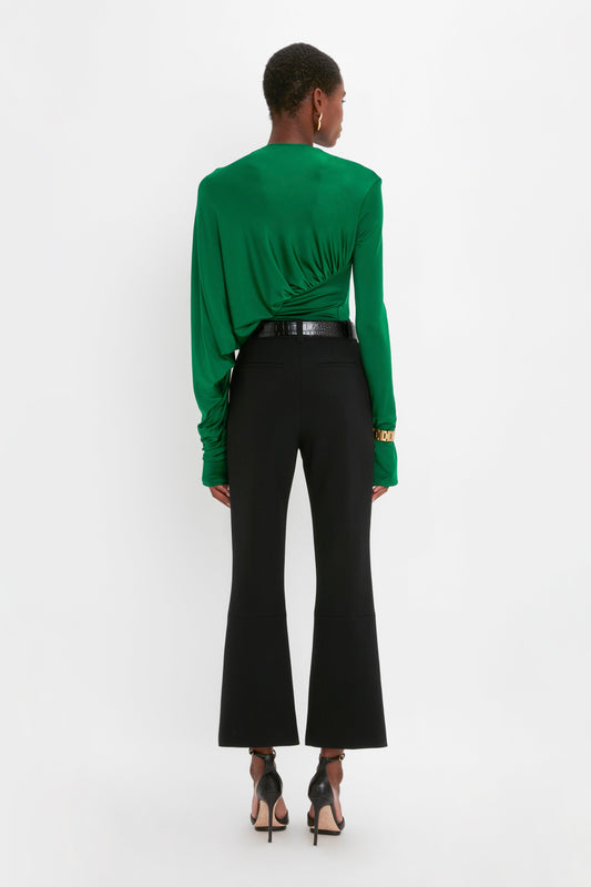 A person stands facing away from the camera, wearing a green long-sleeve top, Victoria Beckham Cropped Kick Trouser In Black, and black high-heeled shoes against a plain white background.