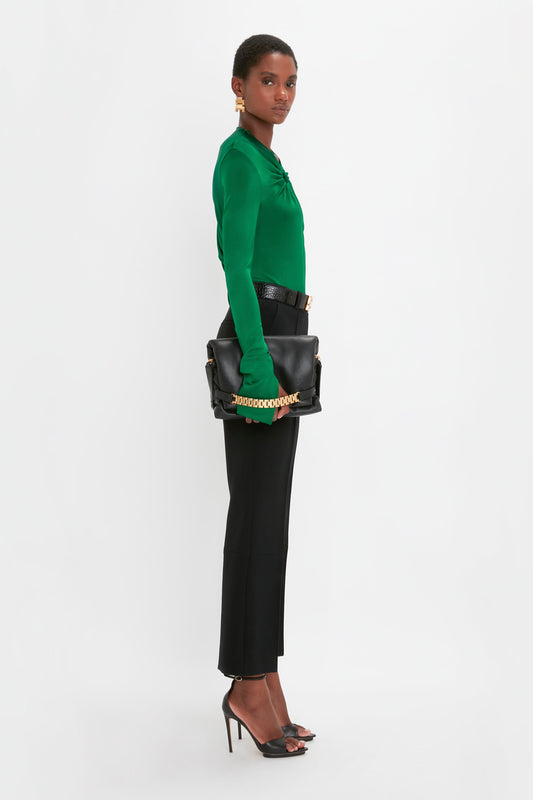 A person stands against a plain background, wearing a green top, Cropped Kick Trouser In Black by Victoria Beckham, high heels, and holding a black clutch bag.