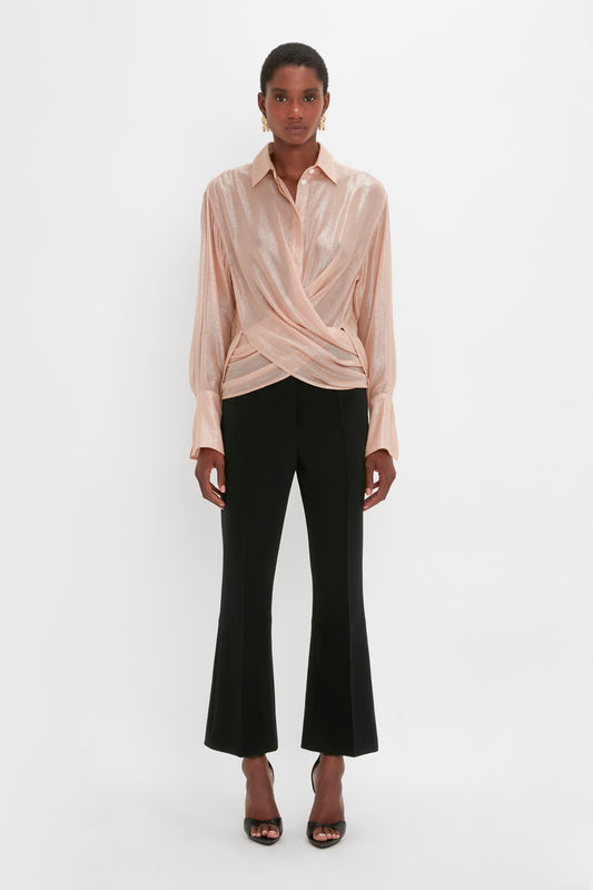 A person stands against a white background wearing the Victoria Beckham Wrap Front Blouse In Rosewater with black flared pants, paired with black high-heeled sandals.