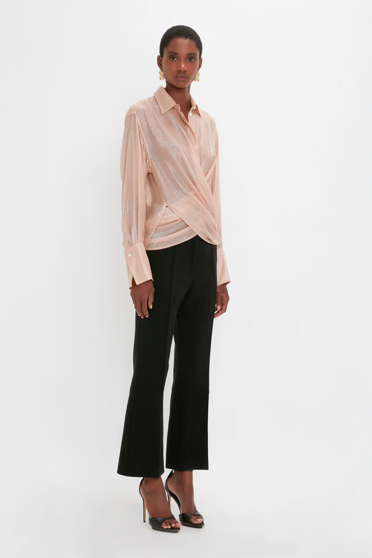 A person poses wearing a Wrap Front Blouse In Rosewater by Victoria Beckham with a draped front and an oversized fit, paired with black wide-leg cropped trousers and high-heeled sandals.