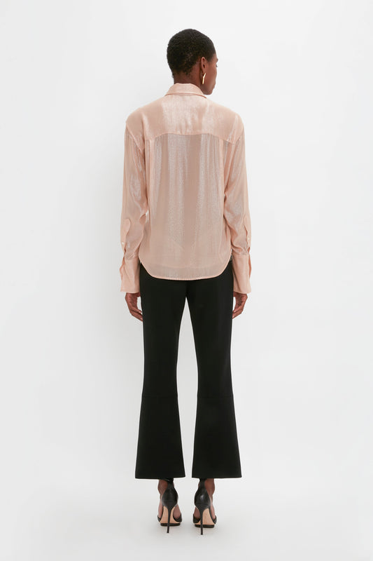 A person stands with their back to the camera, wearing a Victoria Beckham Wrap Front Blouse In Rosewater with an oversized fit, black wide-leg pants, and black high-heeled shoes against a plain white background.