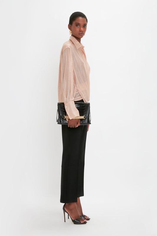 A person stands against a white background, wearing an oversized Victoria Beckham Wrap Front Blouse In Rosewater, black cropped pants, and black heeled sandals, holding a black clutch bag.
