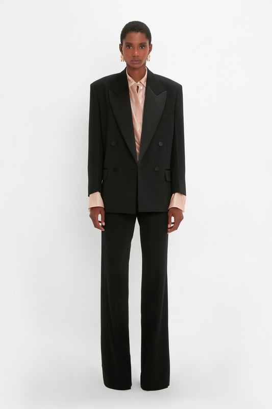 A person is standing against a white background, wearing an oversized black Satin Lapel Tuxedo Jacket in Black by Victoria Beckham, black trousers, and a beige shirt. They have short hair and are wearing earrings.
