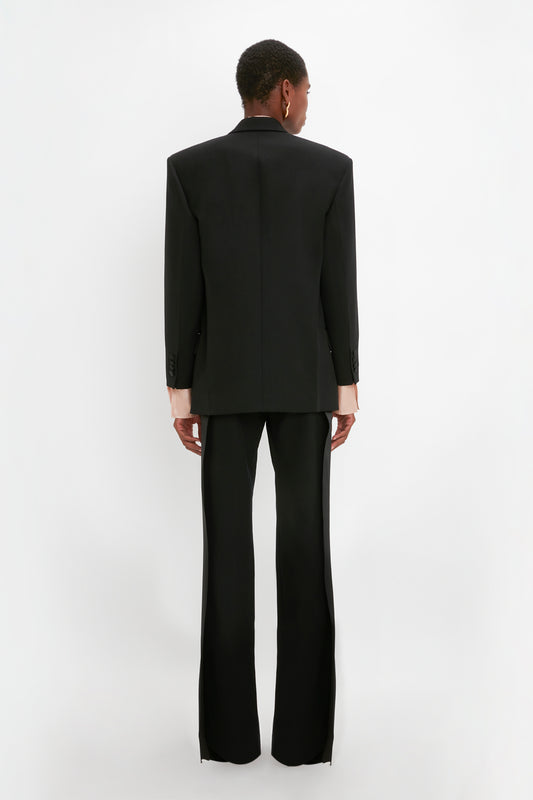 Person standing with their back to the camera, wearing an oversized black Satin Lapel Tuxedo Jacket in Black by Victoria Beckham and flare pants, against a white background.