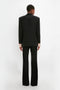 Person standing with their back to the camera, wearing an oversized black Satin Lapel Tuxedo Jacket in Black by Victoria Beckham and flare pants, against a white background.