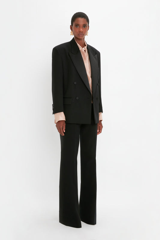 Person standing against a white background wearing an oversized black Satin Lapel Tuxedo Jacket in Black by Victoria Beckham, matching wide-leg trousers, satin gold earrings, and a light pink blouse.