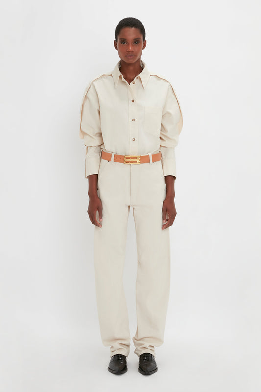A person stands against a white background wearing a Victoria Beckham Oversized Pleat Detail Denim Shirt in Ecru with puffed sleeves, beige trousers, a tan belt, and black shoes.