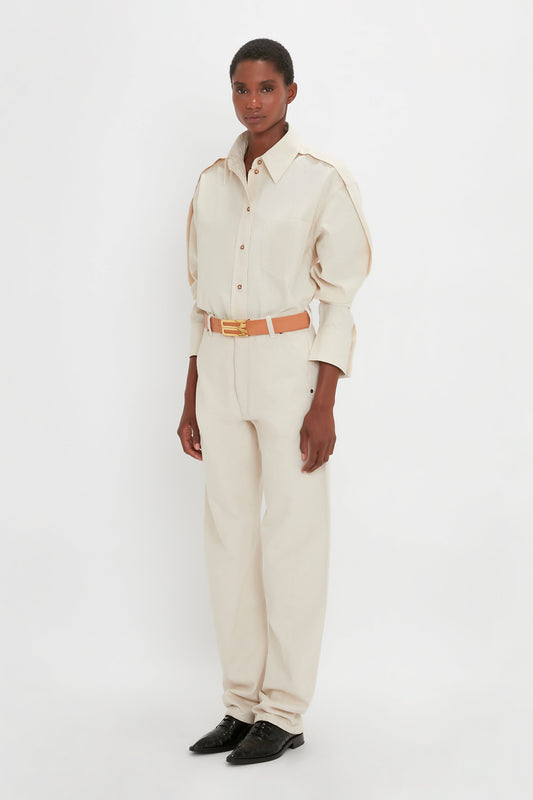 A person stands against a white background, wearing an oversized fit **Oversized Pleat Detail Denim Shirt in Ecru** by **Victoria Beckham**, matching trousers, a tan belt, and black shoes.