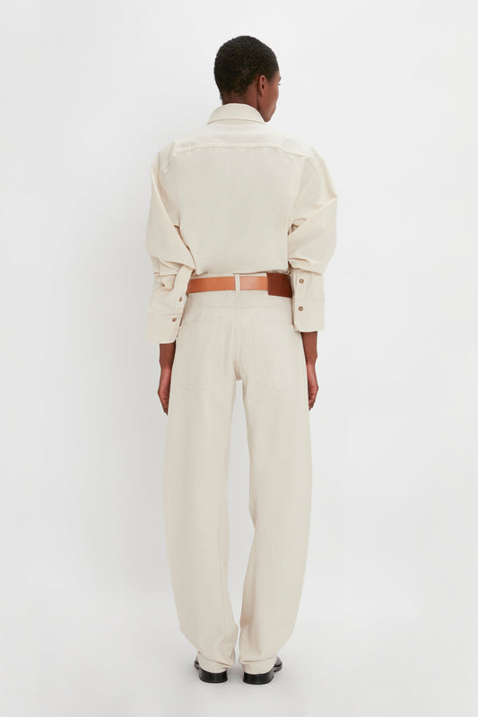 Back view of a person wearing a Relaxed Fit Low-Rise Jean in Ecru by Victoria Beckham with a cream-colored button-up shirt made from breathable cotton and a tan belt, standing against a plain white background.