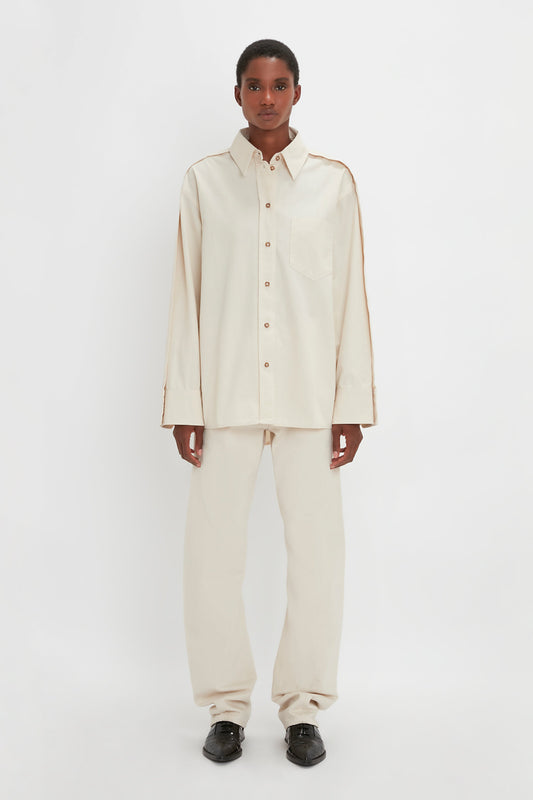 A person stands against a plain white background wearing a Victoria Beckham Oversized Pleat Detail Denim Shirt in Ecru and matching pants with black shoes.