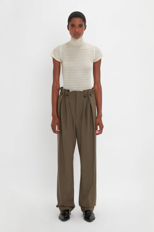 A person stands against a plain white background, wearing a white, short-sleeved turtleneck and high-waisted, wide-leg Gathered Waist Utility Trouser In Oregano by Victoria Beckham paired with black shoes.
