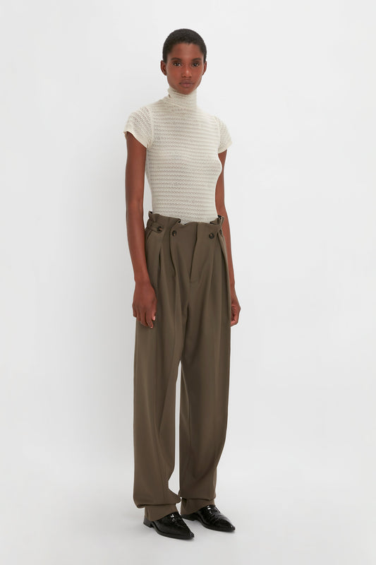 A person is standing against a white background, wearing a white turtleneck top and Victoria Beckham Gathered Waist Utility Trouser in Oregano, paired with black shoes.