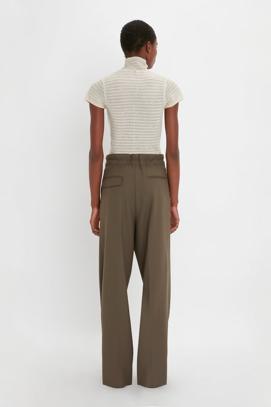 A person with short hair stands with their back facing the camera, wearing a short-sleeved white turtleneck top and loose-fitting Gathered Waist Utility Trouser In Oregano by Victoria Beckham.
