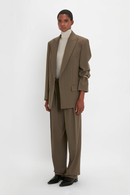 A person stands against a white background wearing an oversized brown suit with a Peak Lapel Jacket In Oregano by Victoria Beckham, a light-colored turtleneck sweater, and black shoes.