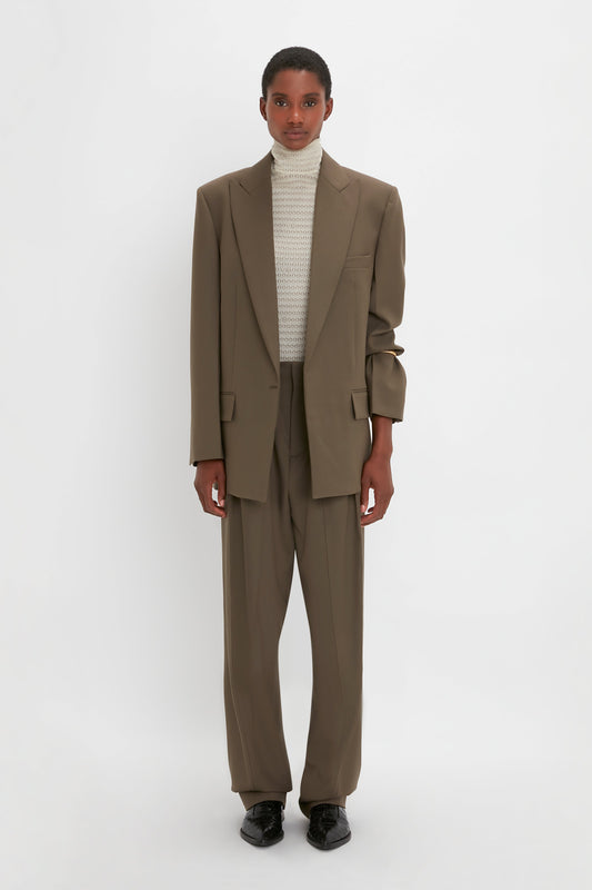 A person stands against a white background wearing an oversized silhouette of a brown suit, highlighted by the Victoria Beckham Peak Lapel Jacket In Oregano paired with a high-neck white knit top and black shoes, capturing a contemporary aesthetic.