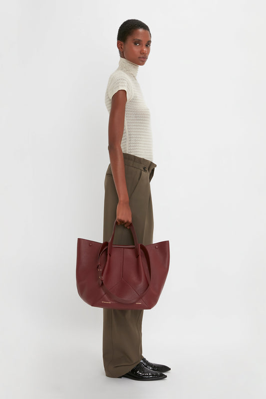 Person standing sideways wearing a cream turtleneck, Victoria Beckham Gathered Waist Utility Trouser In Oregano, black shoes, and holding a large maroon handbag against a plain white background.