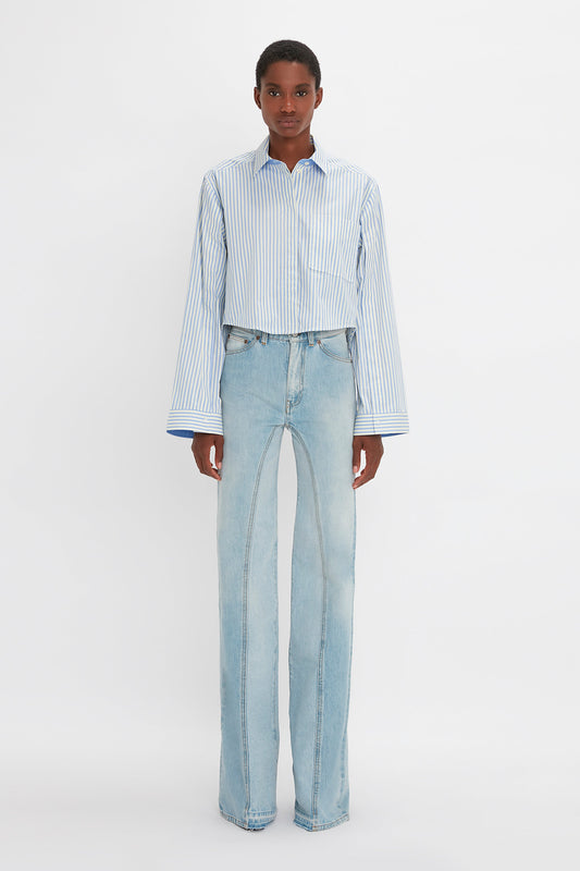 Individual wearing a Victoria Beckham Button Detail Cropped Shirt In Chamomile Blue Stripe and high-waisted, light-wash flared jeans, boasting a feminine silhouette while standing in front of a plain white background.