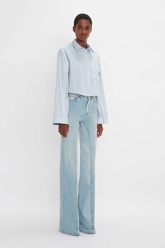 A person is standing in front of a plain white background, wearing a Button Detail Cropped Shirt In Chamomile Blue Stripe by Victoria Beckham and high-waisted, wide-leg light blue jeans, showcasing a versatile styling that blends both feminine silhouette and masculine tailoring.
