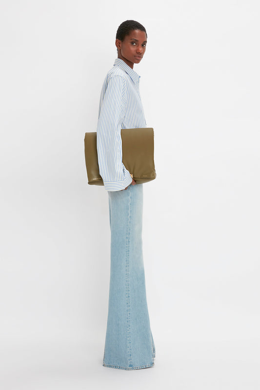 A person in a **Victoria Beckham Button Detail Cropped Shirt In Chamomile Blue Stripe** and light blue high-waisted jeans, showcasing a feminine silhouette, holds a tan clutch against a plain white background.