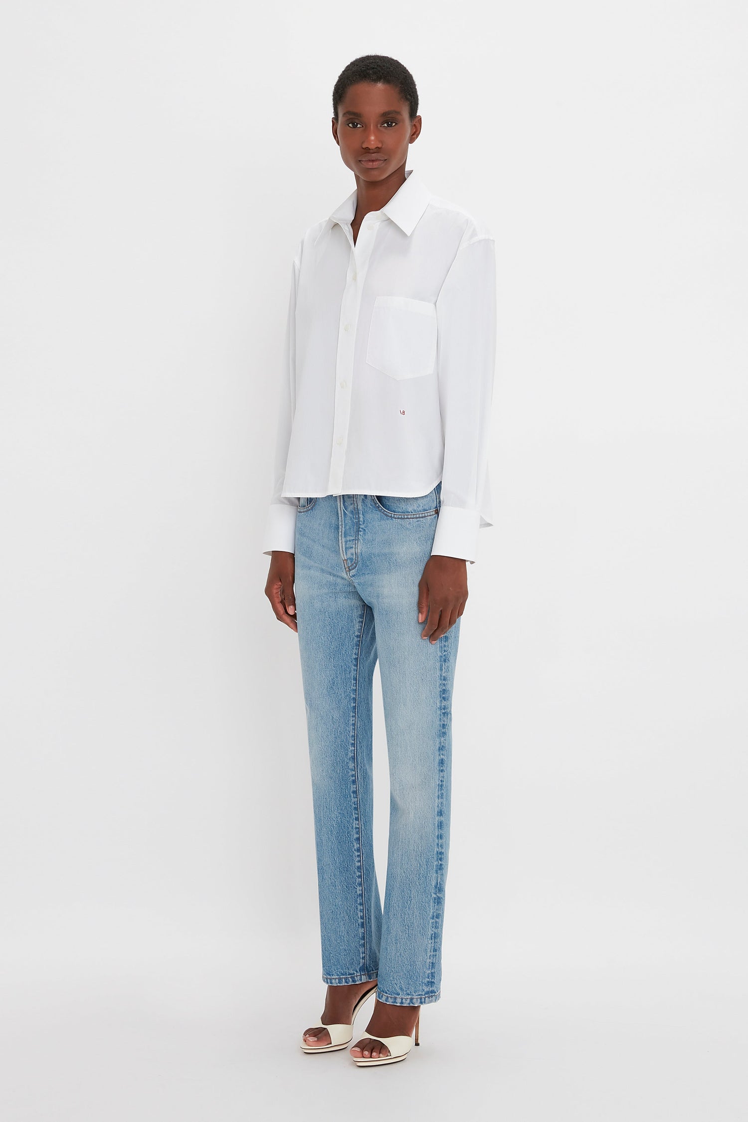 A woman in a classic white Cropped Long Sleeve Shirt In White by Victoria Beckham monogram and blue jeans stands facing the camera against a plain white background.