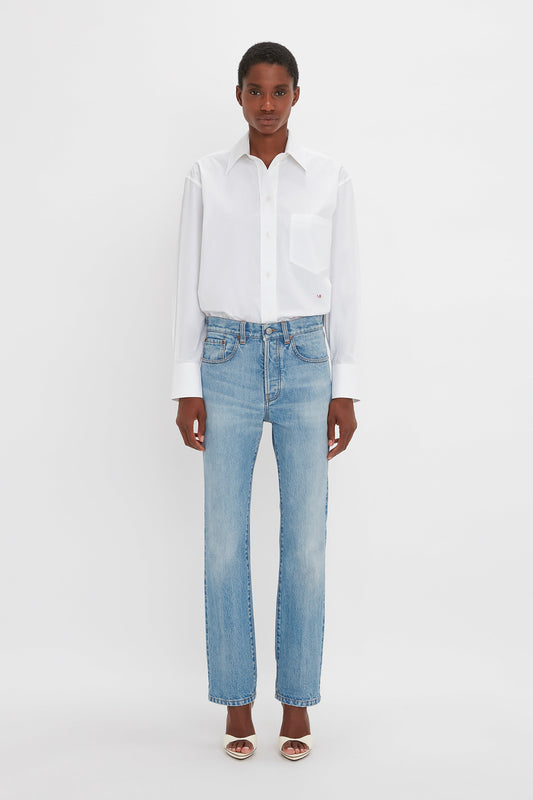 A person stands against a plain background, wearing a white button-up shirt, Victoria Beckham Victoria Mid-Rise Jean In Light Blue, and beige sandals.