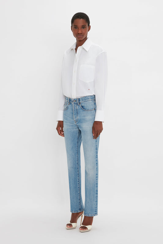 A person stands against a plain white background, wearing a white button-up shirt, Victoria Mid-Rise Jean In Light Blue by Victoria Beckham, and white open-toe heels.