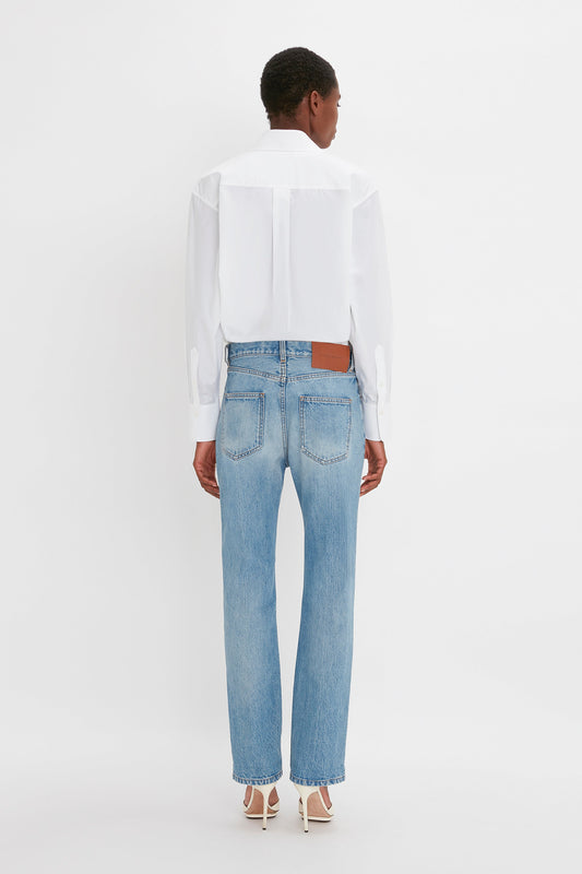 Person standing facing away, wearing a white long-sleeve shirt, Victoria Mid-Rise Jean In Light Blue by Victoria Beckham, and white shoes, against a plain white background.