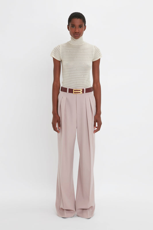 A person stands against a white background wearing a short-sleeved, high-necked, cream-colored Victoria Beckham Polo Neck Knitted T-Shirt In Cream paired with wide-leg, high-waisted, light pink trousers and a brown belt with a gold buckle.