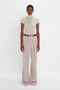 A person stands against a white background wearing a short-sleeved, high-necked, cream-colored Victoria Beckham Polo Neck Knitted T-Shirt In Cream paired with wide-leg, high-waisted, light pink trousers and a brown belt with a gold buckle.