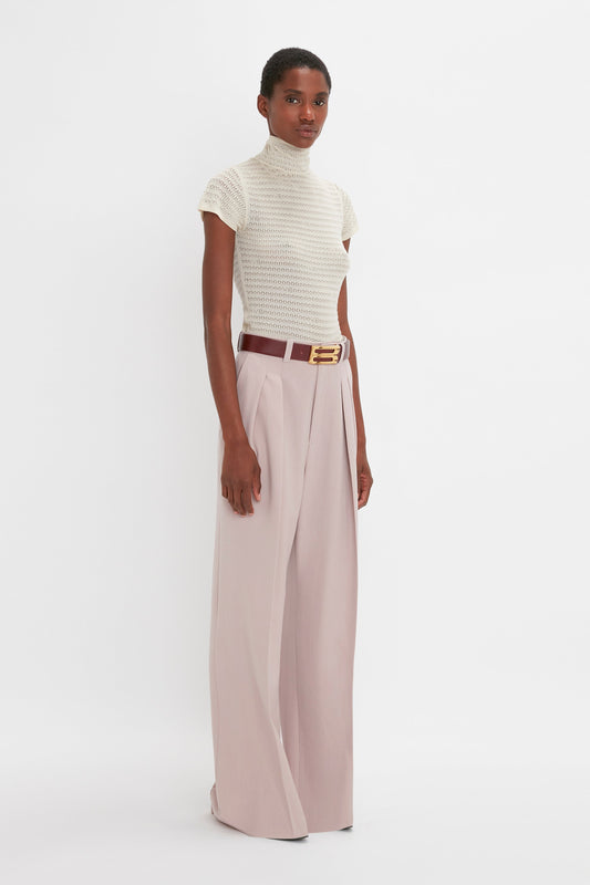 A person stands in a neutral pose wearing a Polo Neck Knitted T-Shirt In Cream by Victoria Beckham and high-waisted wide-leg pink trousers with a brown belt.