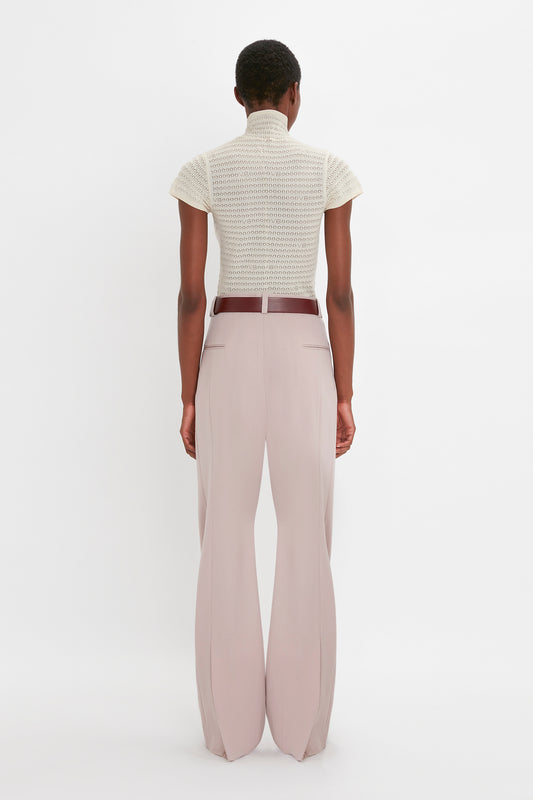 A person is standing with their back to the camera, wearing a beige Polo Neck Knitted T-Shirt In Cream by Victoria Beckham and light pink wide-leg pants with a brown belt against a plain white background. The outfit features trans-seasonal appeal, perfect for mixing and matching throughout the year.