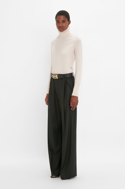 A woman standing in a studio, wearing a white Victoria Beckham lambswool polo neck jumper in ivory and black trousers with a gold belt.
