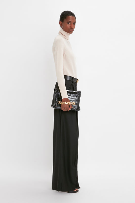 Woman in a chic Victoria Beckham ivory polo neck jumper and black trousers holding a studded black handbag, standing against a plain white background.
