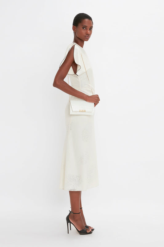 Person in a sleeveless white dress and black heels stands sideways, holding an Exclusive Wallet On Chain In Ivory Croc-Effect Leather by Victoria Beckham.