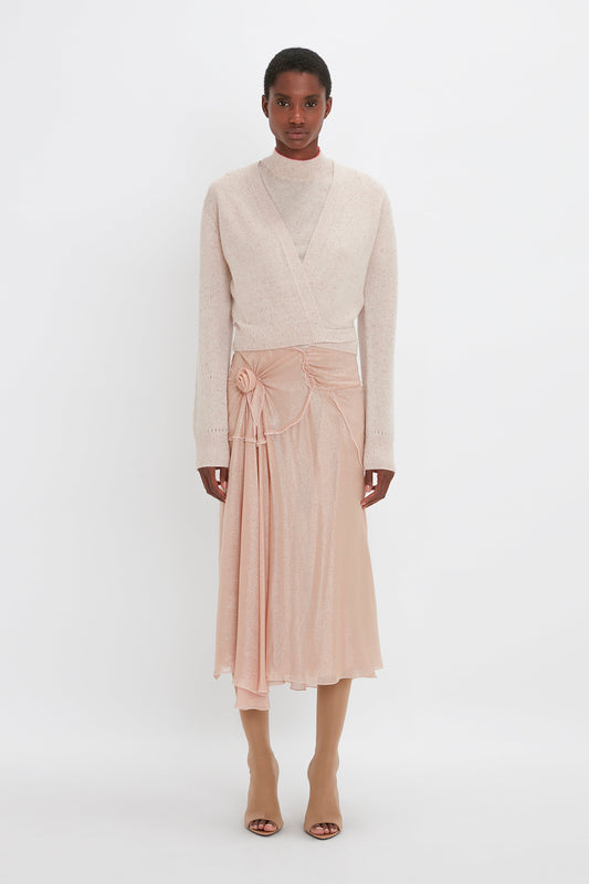 A person stands against a plain white background wearing a light-colored wrap sweater, a high-neck top, a Flower Detail Cami Skirt In Rosewater by Victoria Beckham, and beige boots.