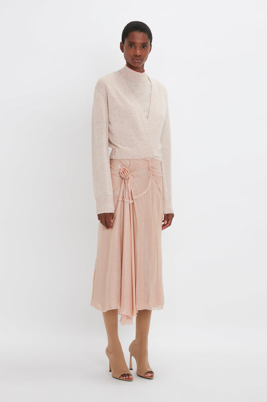 A person stands against a white background, wearing a cream sweater, a light pink Flower Detail Cami Skirt In Rosewater by Victoria Beckham, and tan open-toe boots.